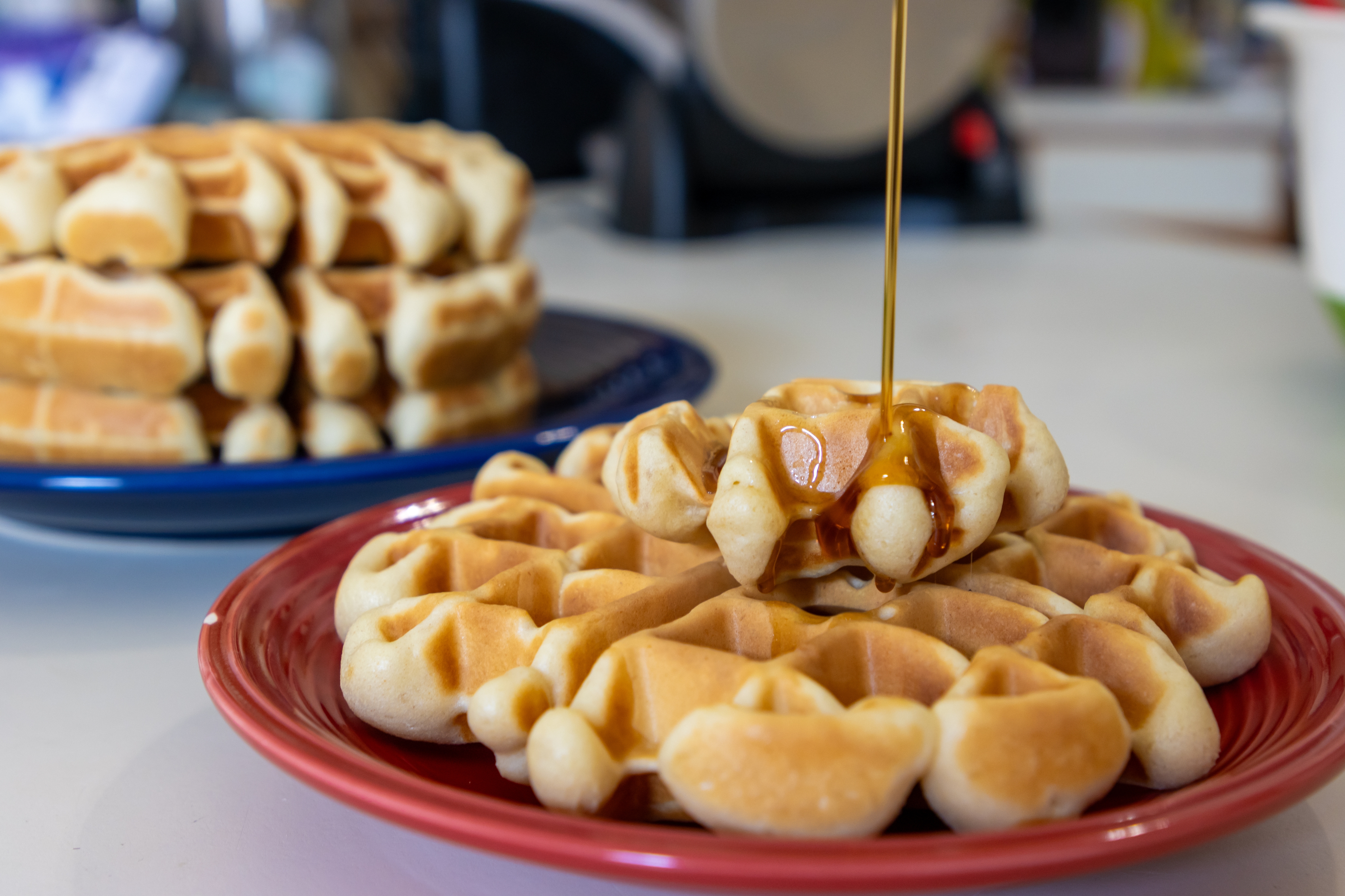 Syrup being poured over a stack of waffles on a red plate, with more waffles on a blue plate in the background