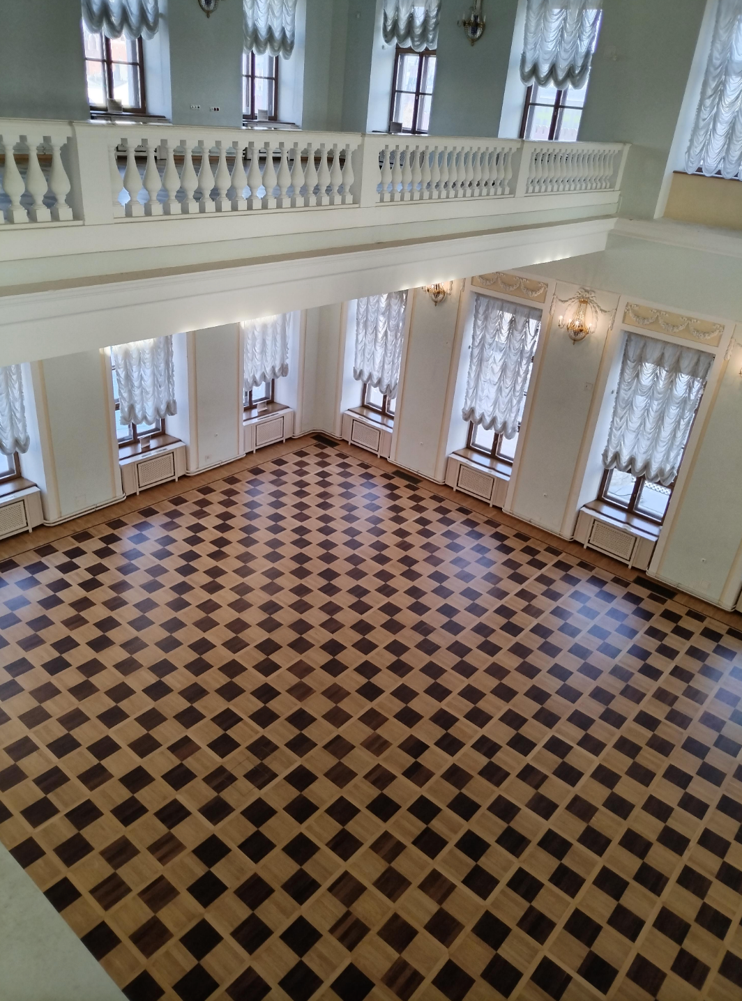Spacious ballroom with large windows, elegant drapes, and a checkered wooden floor, viewed from above