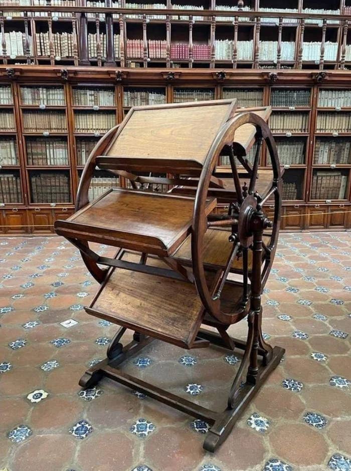 A vintage rotating book stand with multiple shelves, set in a large library with many bookshelves in the background