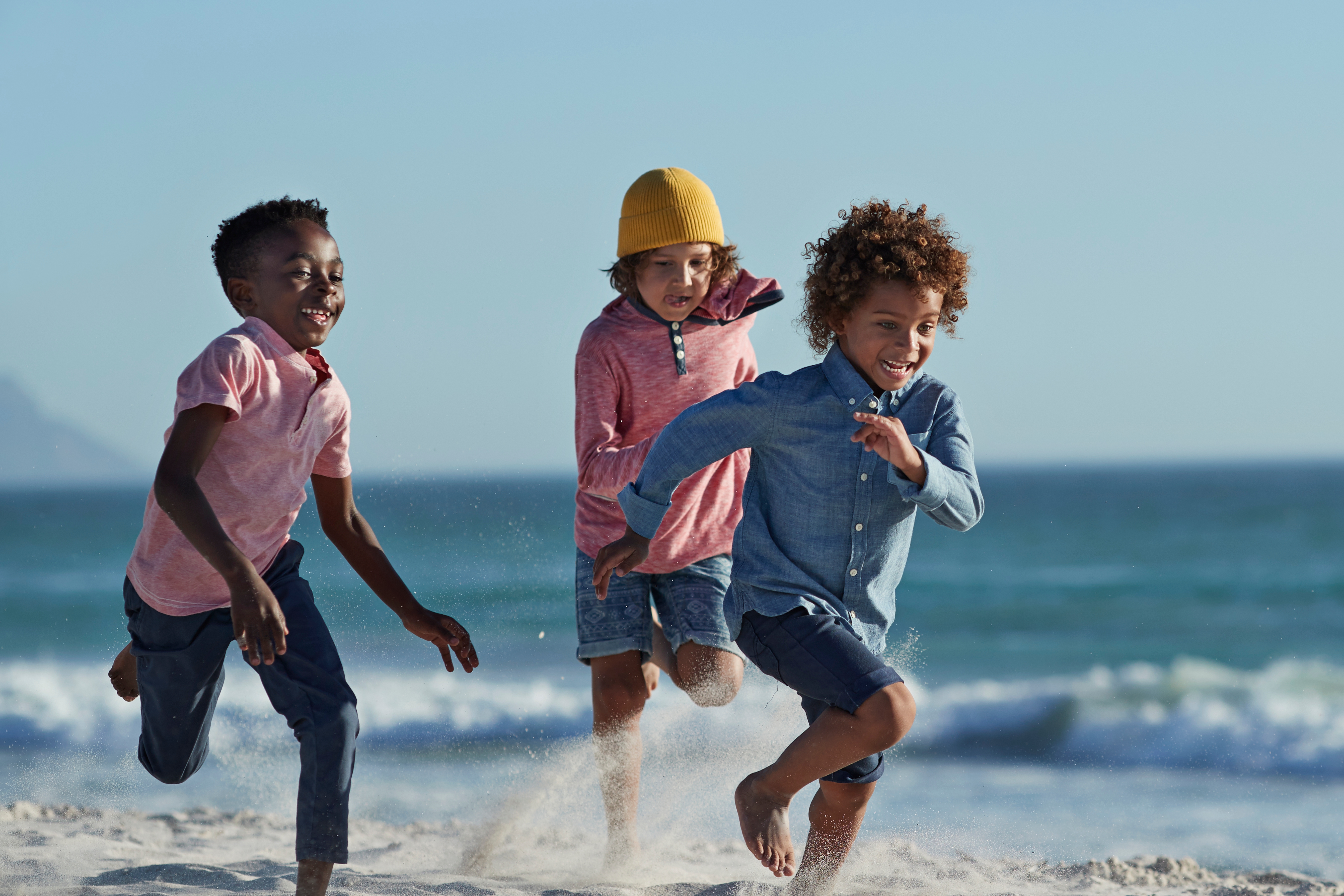Three children joyfully run on a sandy beach near the ocean, their energy and carefree spirit captured as waves roll in the background