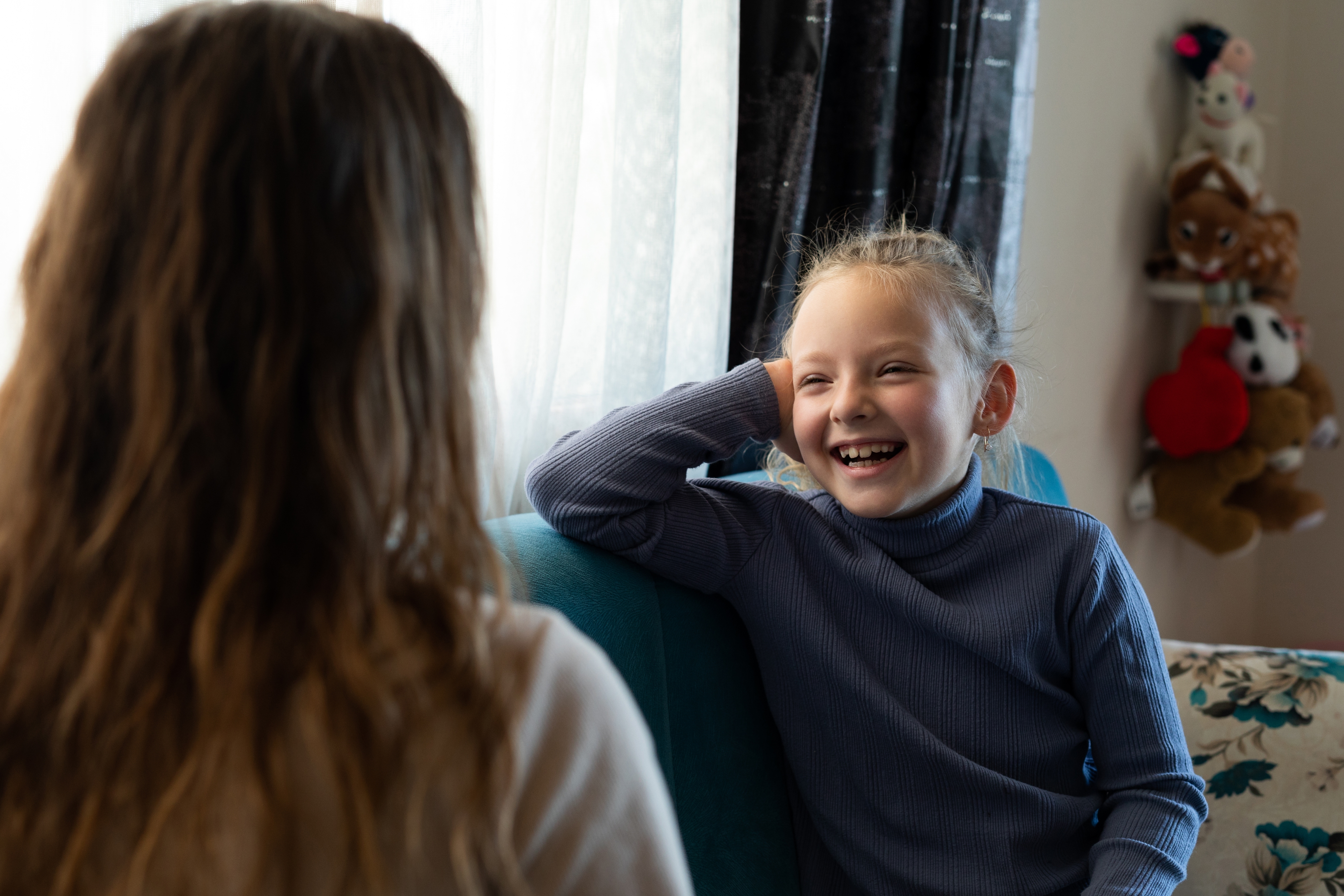 Child laughing while talking to an adult on a couch at home, stuffed toys visible in the background