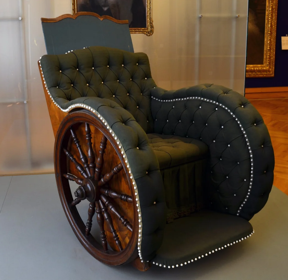 Antique wheelchair with tufted upholstery, wooden wheels, and nailhead trim, displayed in a museum setting