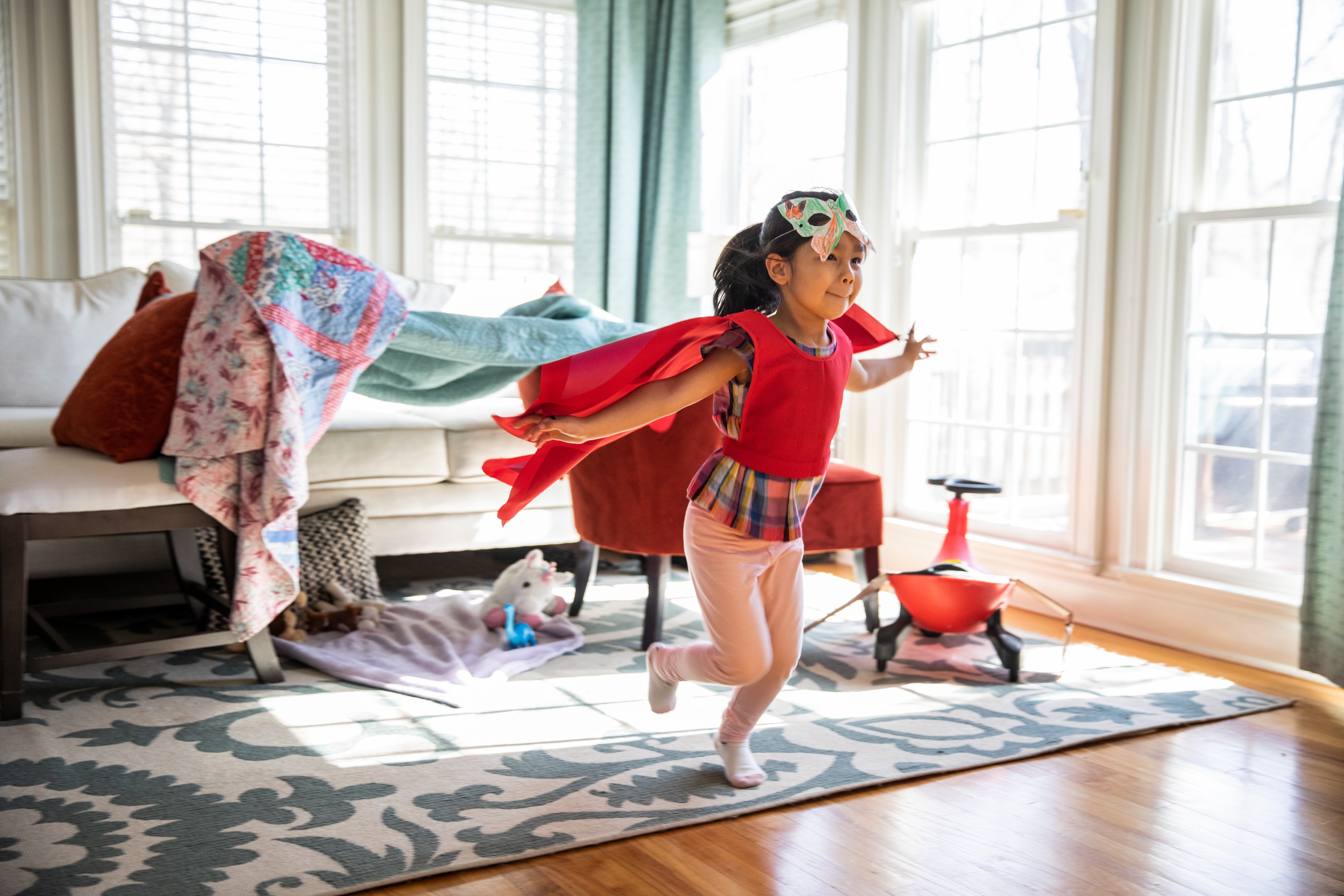 Child playing superhero with a cape and mask in a sunlit living room, embodying imagination and play