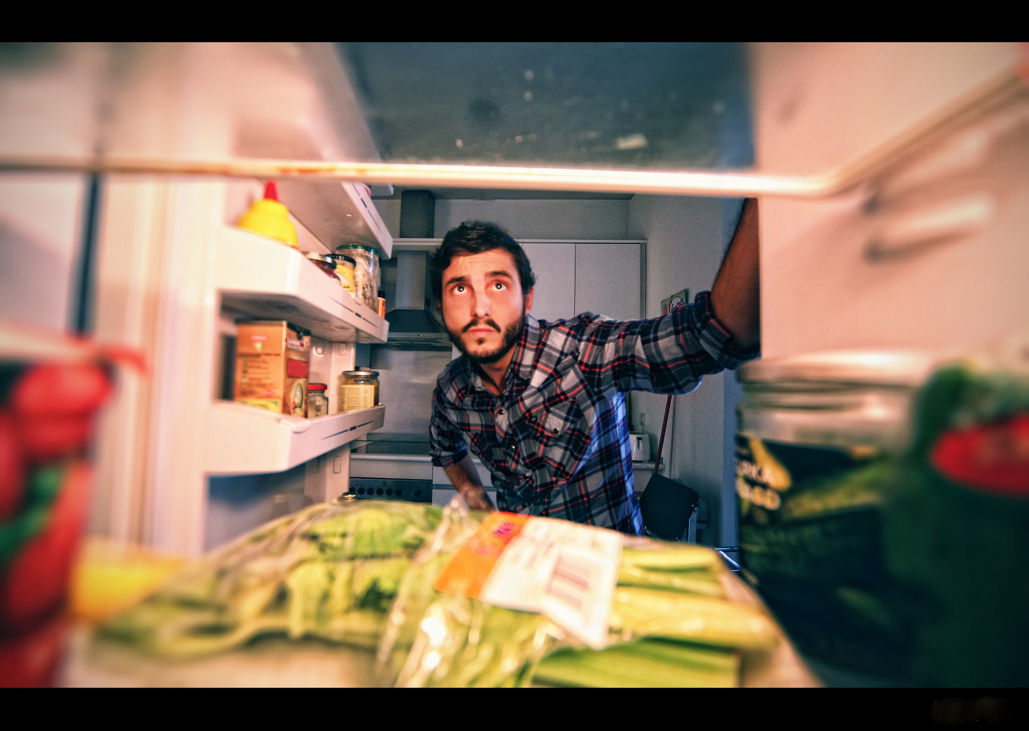 Man looks inside an open fridge filled with various food items. He appears thoughtful, scanning the contents. Kitchen background visible