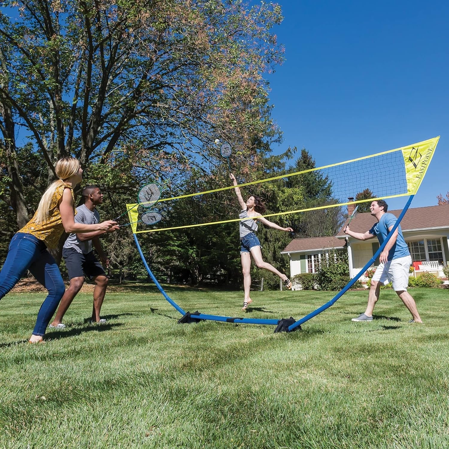Four people playing badminton in a backyard, using a portable net set. Two teams are actively engaged in the game