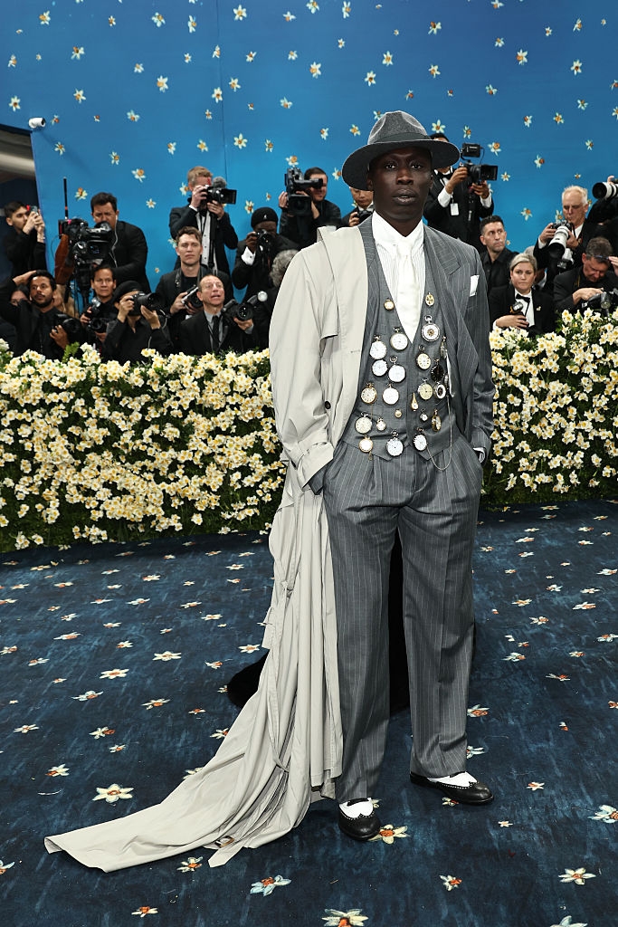 Person on red carpet in pin-adorned gray suit with a long, trailing coat and matching hat, surrounded by photographers and floral backdrop