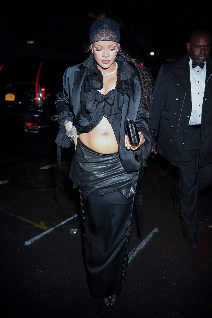 Mujer embarazada con blusa negra con volantes, falda larga y saco, camina en un evento nocturno. Lleva un bolso y está acompañada