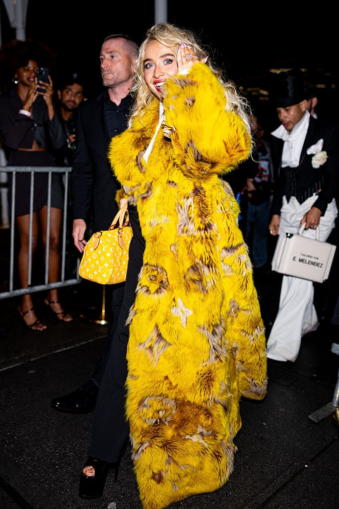 Persona con abrigo llamativo y bolso de mano, saludando mientras camina por un evento nocturno. Otros invitados detrás observan la escena