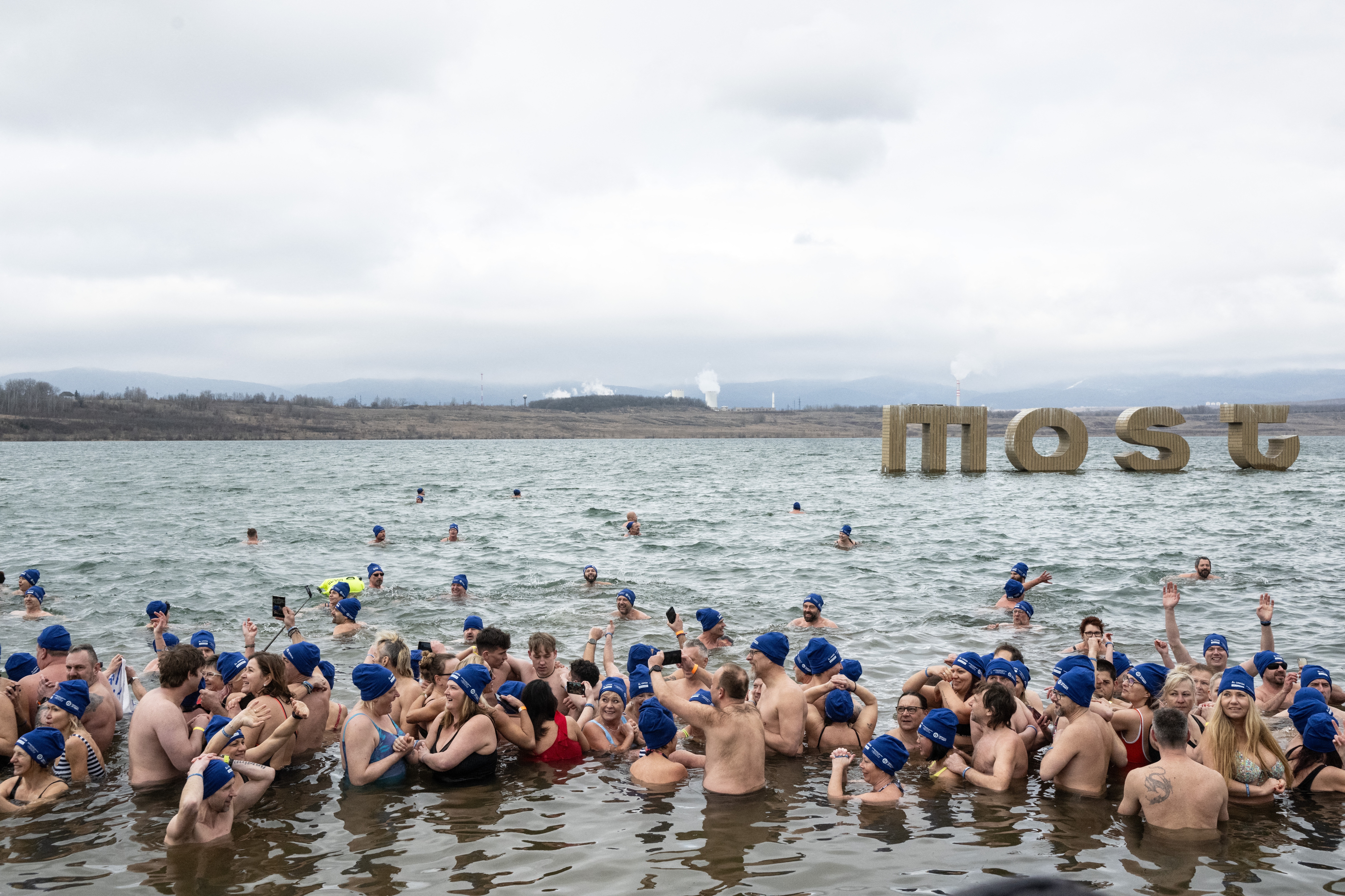 Group of people in swimsuits and blue caps gather in chilly water for a winter swim event, a large &quot;MOST&quot; sign visible in the background