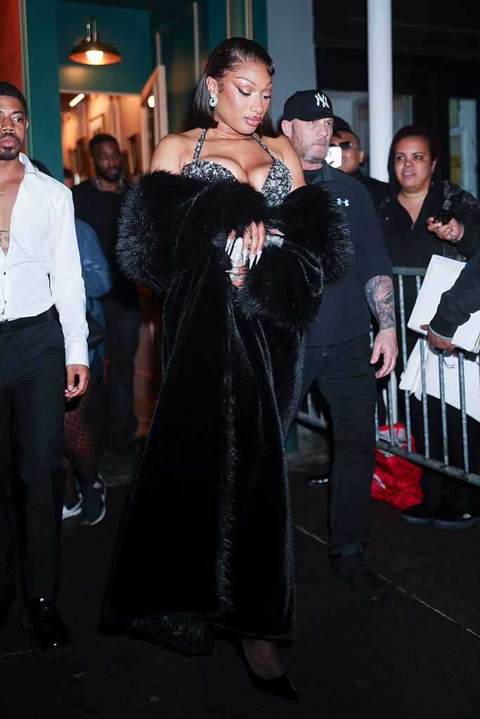 Persona con vestido elegante de gala y abrigo largo, rodeada de gente en una alfombra roja. Viste joyas llamativas. Ambiente de evento nocturno