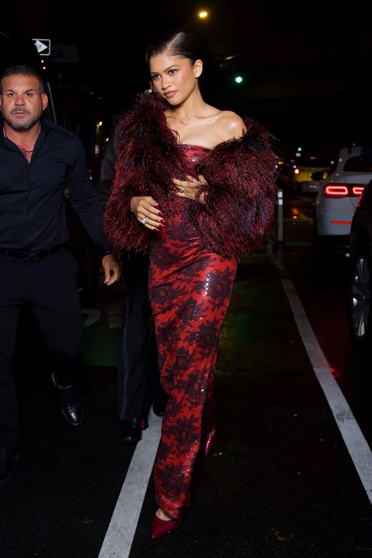 Persona con vestido ajustado y brillante de lentejuelas, envuelta en chal peludo, camina por la calle de noche acompañada por otra persona