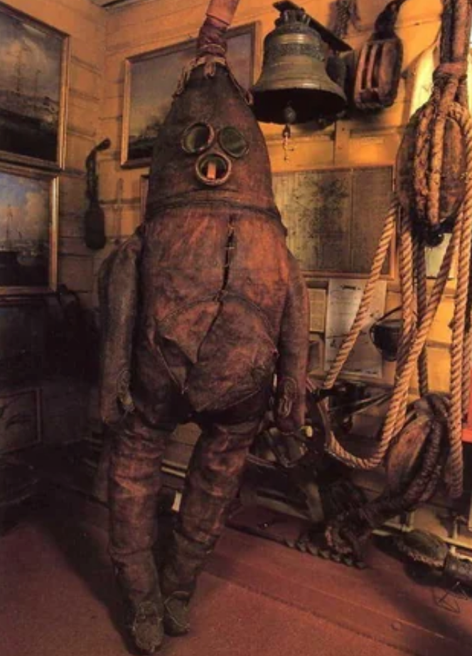 Antique diving suit with a helmet and hose on display in a nautical-themed room with ropes, paintings, and a bell