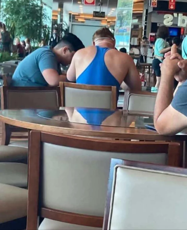 A person is seated at a table in a public area, wearing a sleeveless top; the reflection makes it look like he's wearing a speedo bottom, or a one-piece
