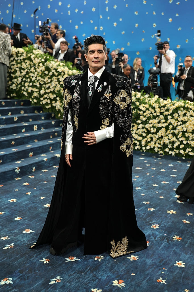 Person in ornate, patterned cape and formal suit on floral carpet at celebrity event, surrounded by photographers