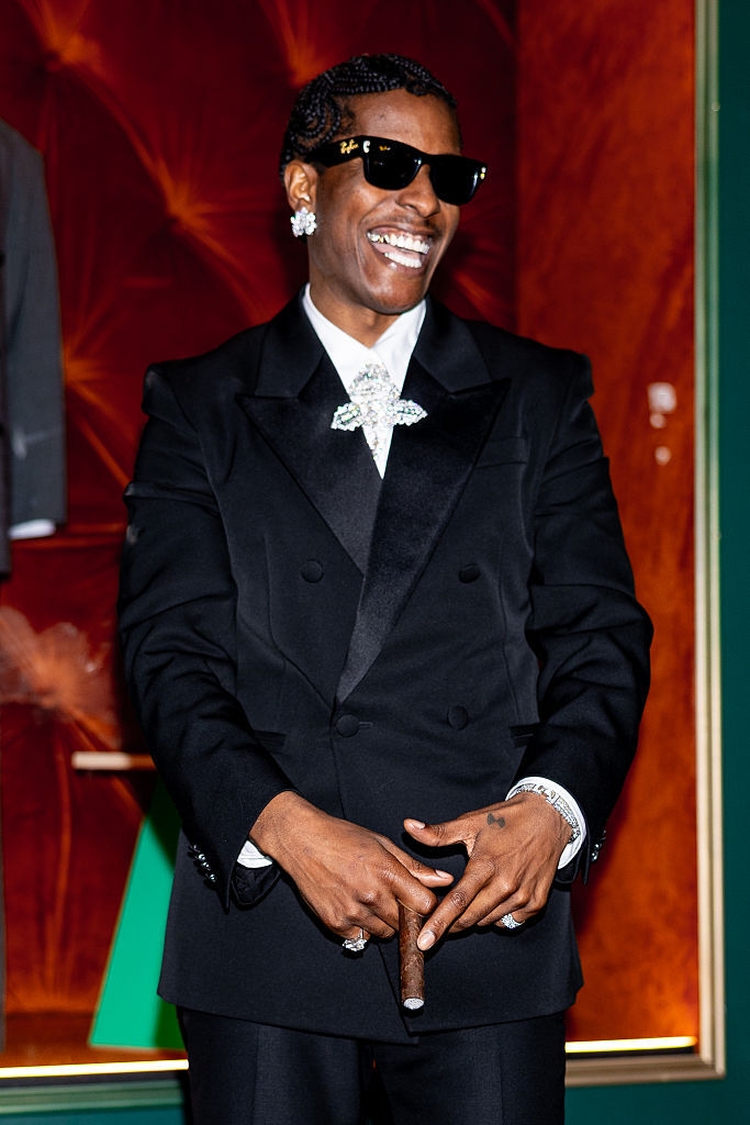 Persona sonriendo con traje elegante, gafas de sol y un puro en la mano. Lleva accesorios brillantes y tiene el cabello peinado hacia atrás