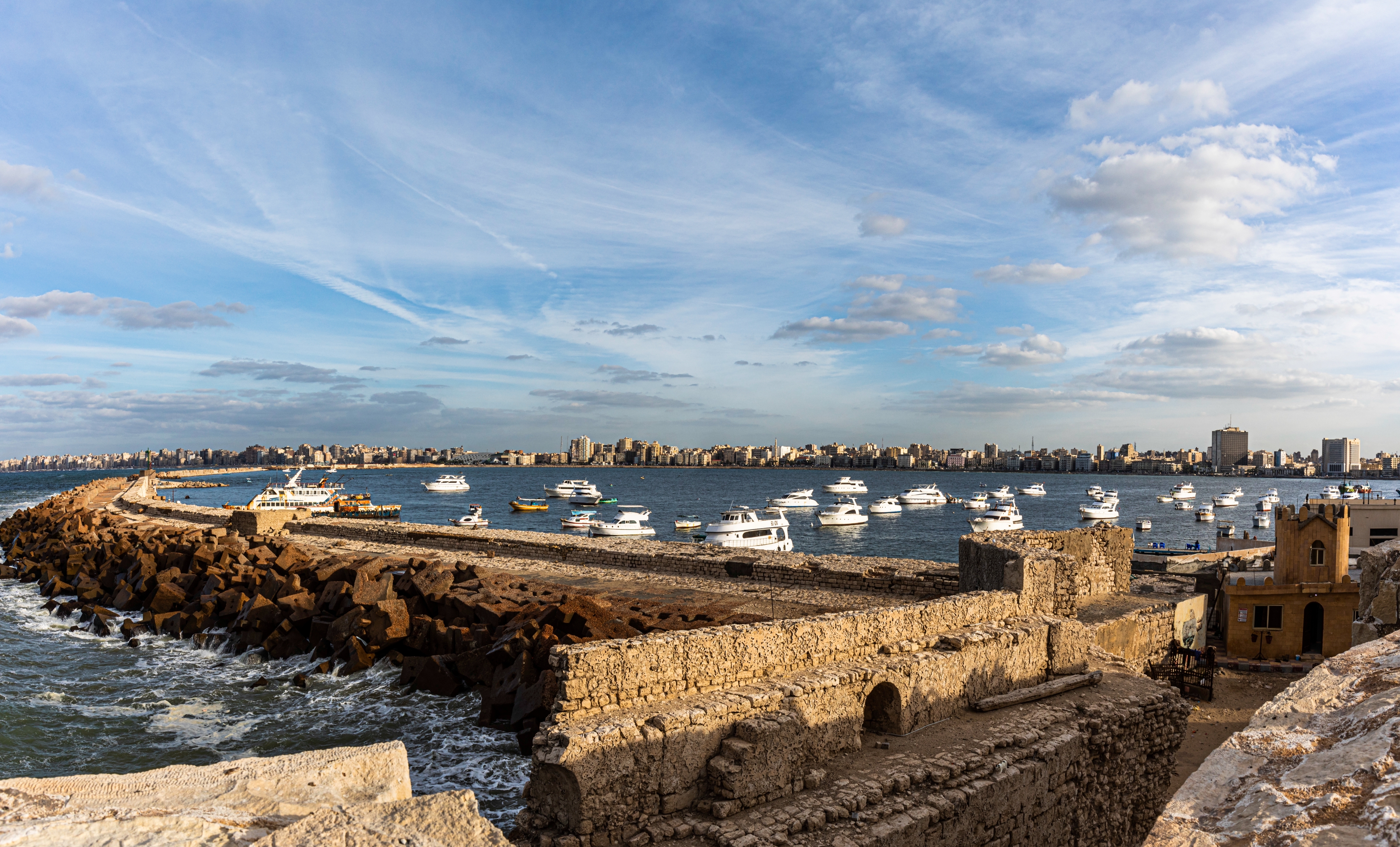Panoramic view of a historic seaside fortress with numerous boats anchored in a calm harbor beneath a wide sky