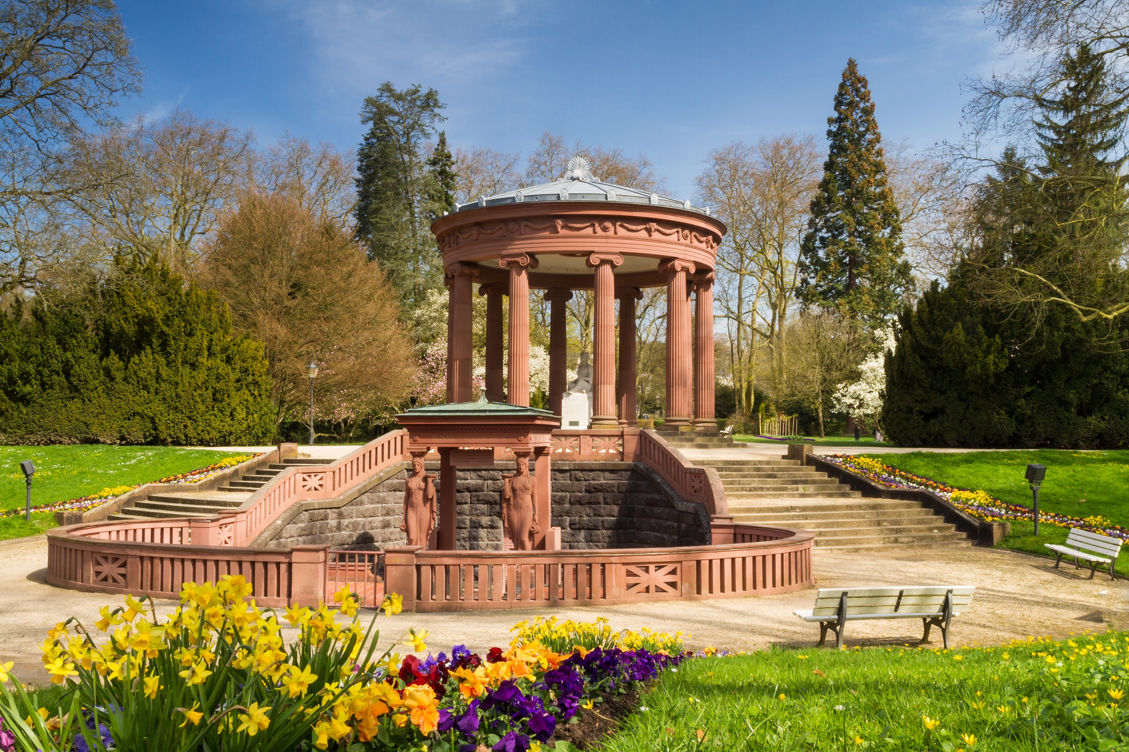 Ornate stone gazebo in a park, surrounded by trees and vibrant flower beds, with benches in the foreground