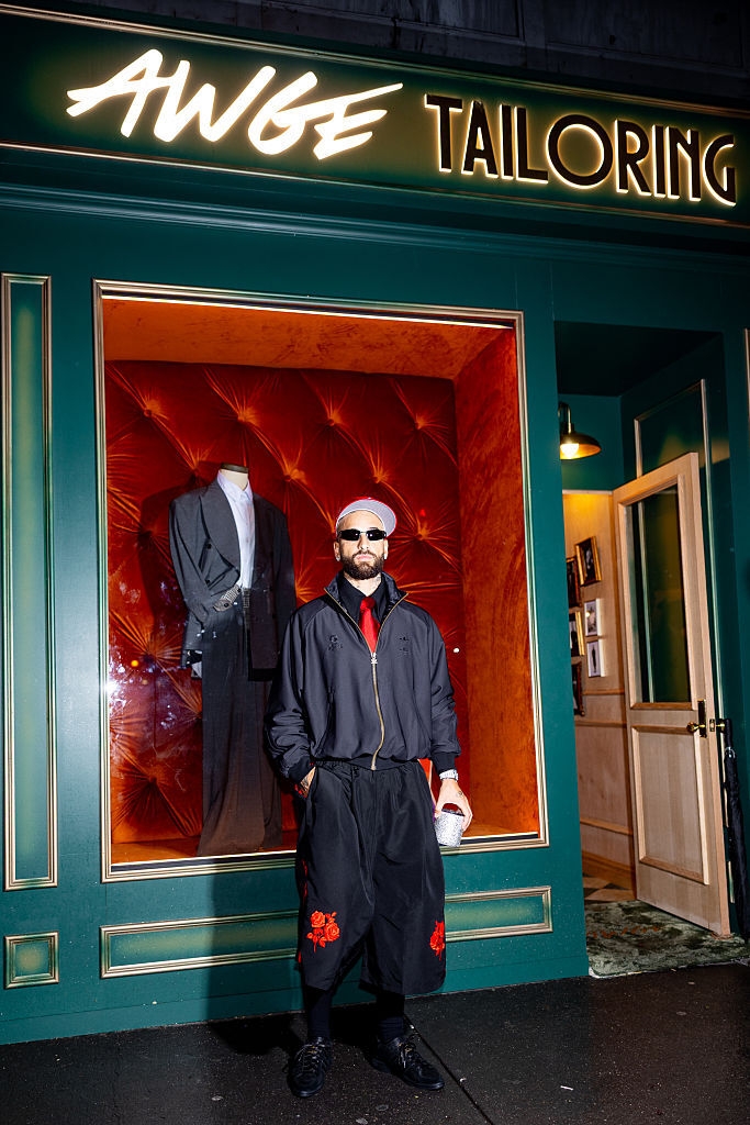 Persona frente a una sastrería, lleva ropa deportiva con chaqueta y pantalones cortos amplios, mientras sostiene una bebida