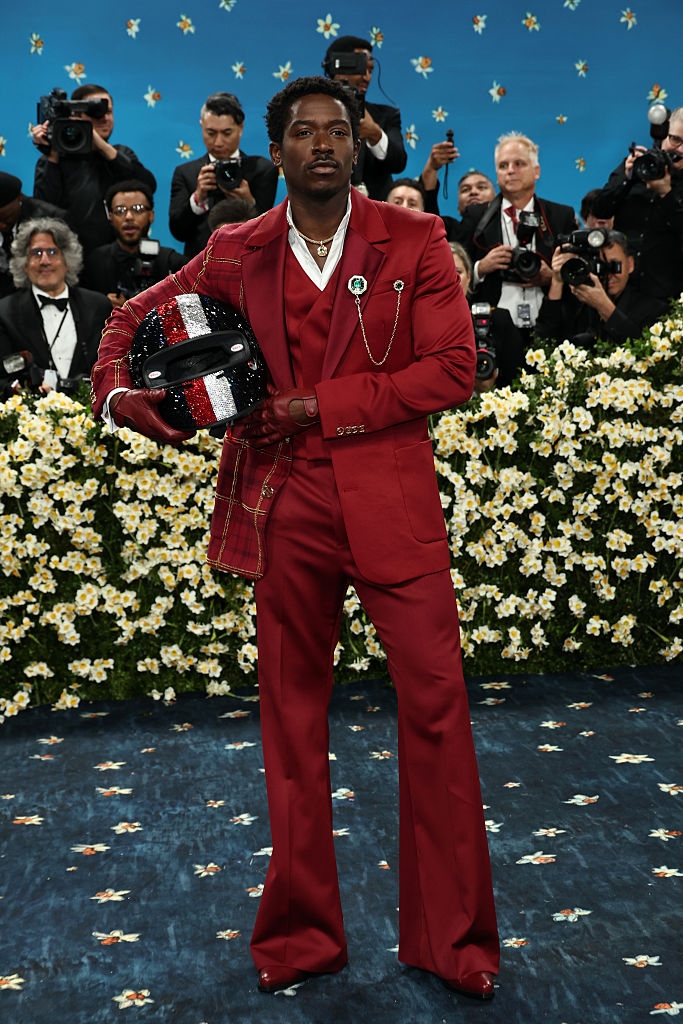 A person in a stylish red suit with a glittering helmet poses confidently on a floral-themed red carpet, surrounded by photographers