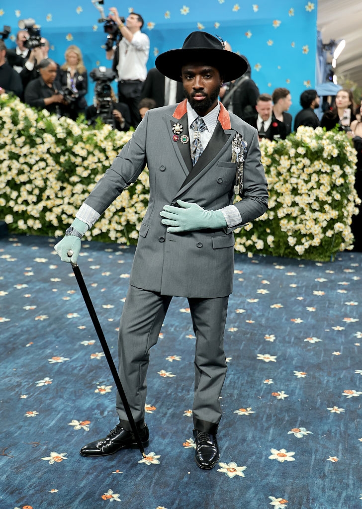 Person in gray suit, gloves, hat, and cane on floral carpet, posing at event. Crowd and photographers in background
