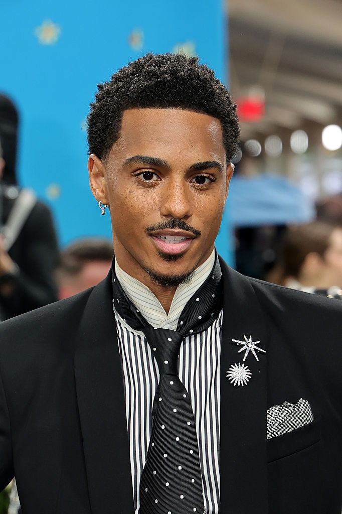 Person in a black suit with striped shirt and polka dot tie, featuring starburst brooch and pocket square, attends a formal event