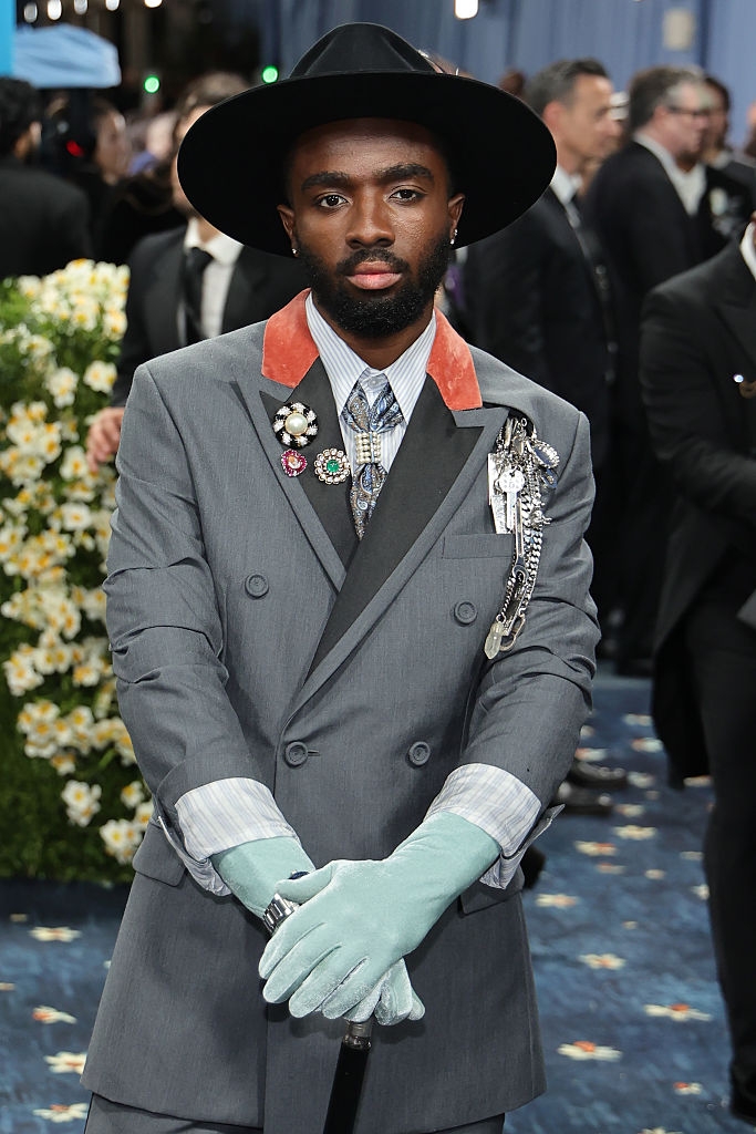 A person on the red carpet wearing a stylish gray suit with embellished lapels, light gloves, and a wide-brimmed hat, holding a cane