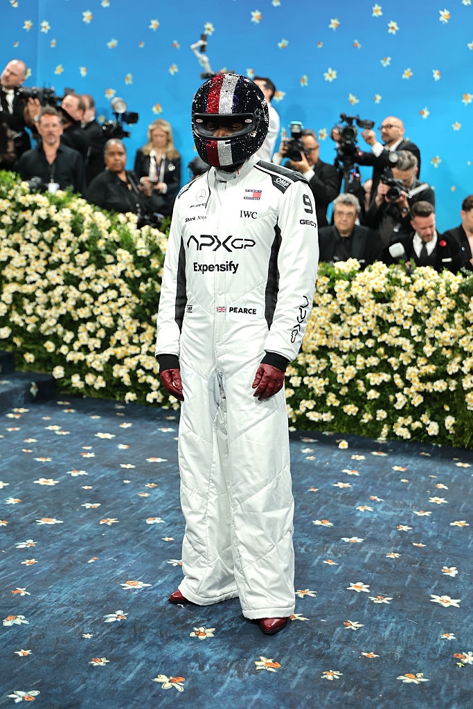 Person in a racing-style jumpsuit and helmet on a blue carpet, surrounded by photographers and floral decorations at a celebrity event