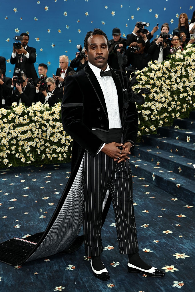 A person stands on a red carpet in a velvet tuxedo with pinstripe pants and a long trailing cape, surrounded by photographers and floral decor