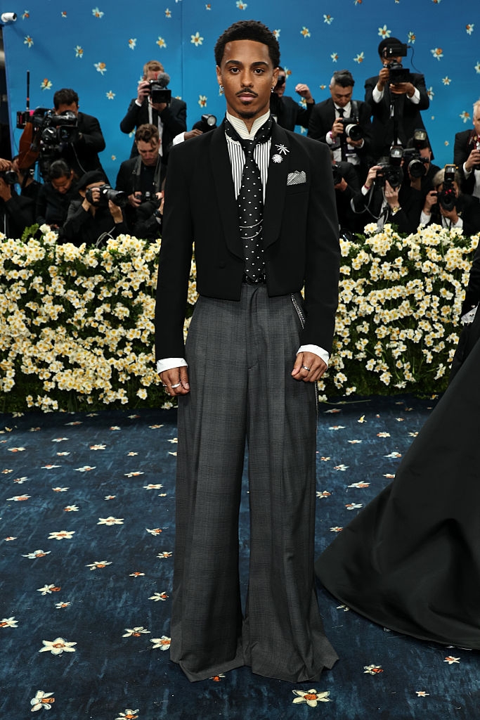 Individual on the red carpet wearing a cropped tuxedo jacket, striped shirt, polka dot tie, and wide-leg gray trousers. Photographers are in the background