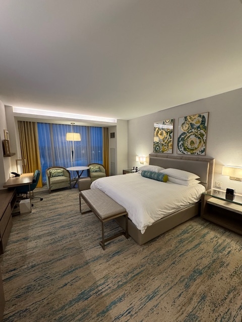 Spacious hotel room with a king-size bed, seating area near large window, desk, and modern decor, ideal for travelers seeking comfort and style