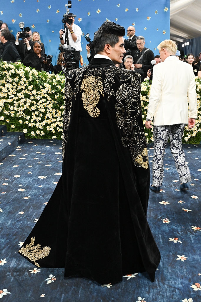 Person in elaborate embroidered cape on red carpet, surrounded by photographers and floral backdrop. Another person in patterned pants walks ahead