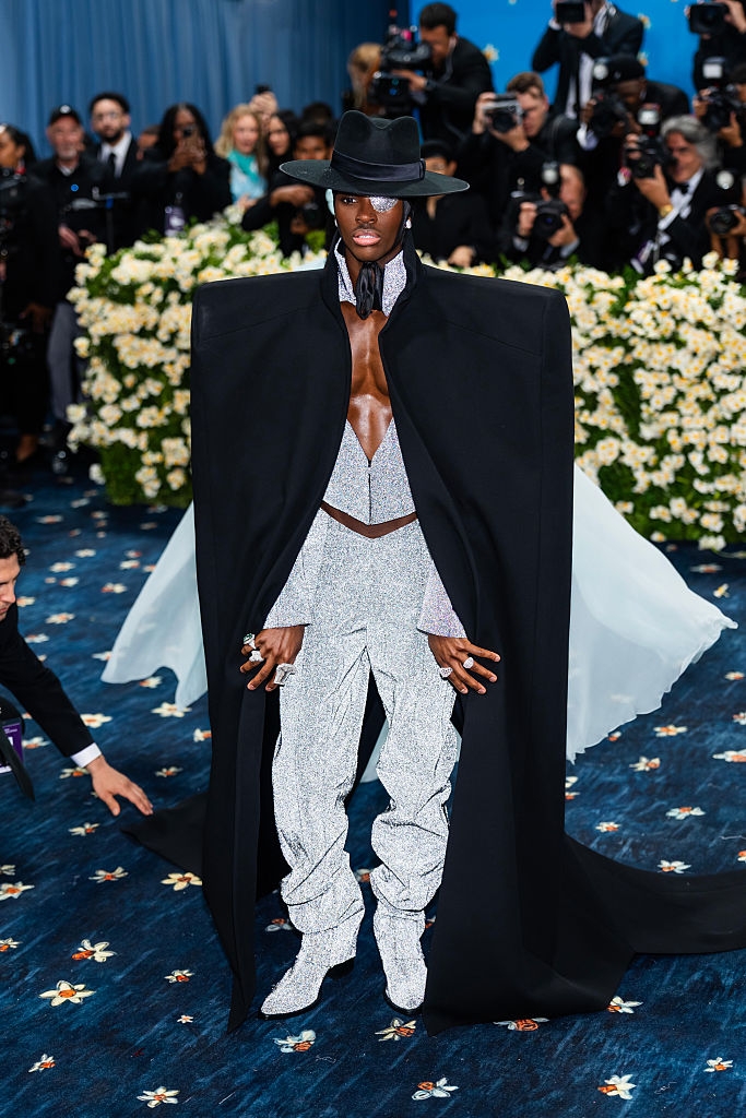 Person in a glittery outfit with a dramatic black cape and hat, posed on a floral-patterned carpet at a celebrity event