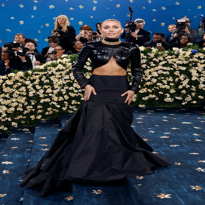 Person in a fitted, long black gown with a high neckline and cutout midriff, posing on a floral-themed red carpet