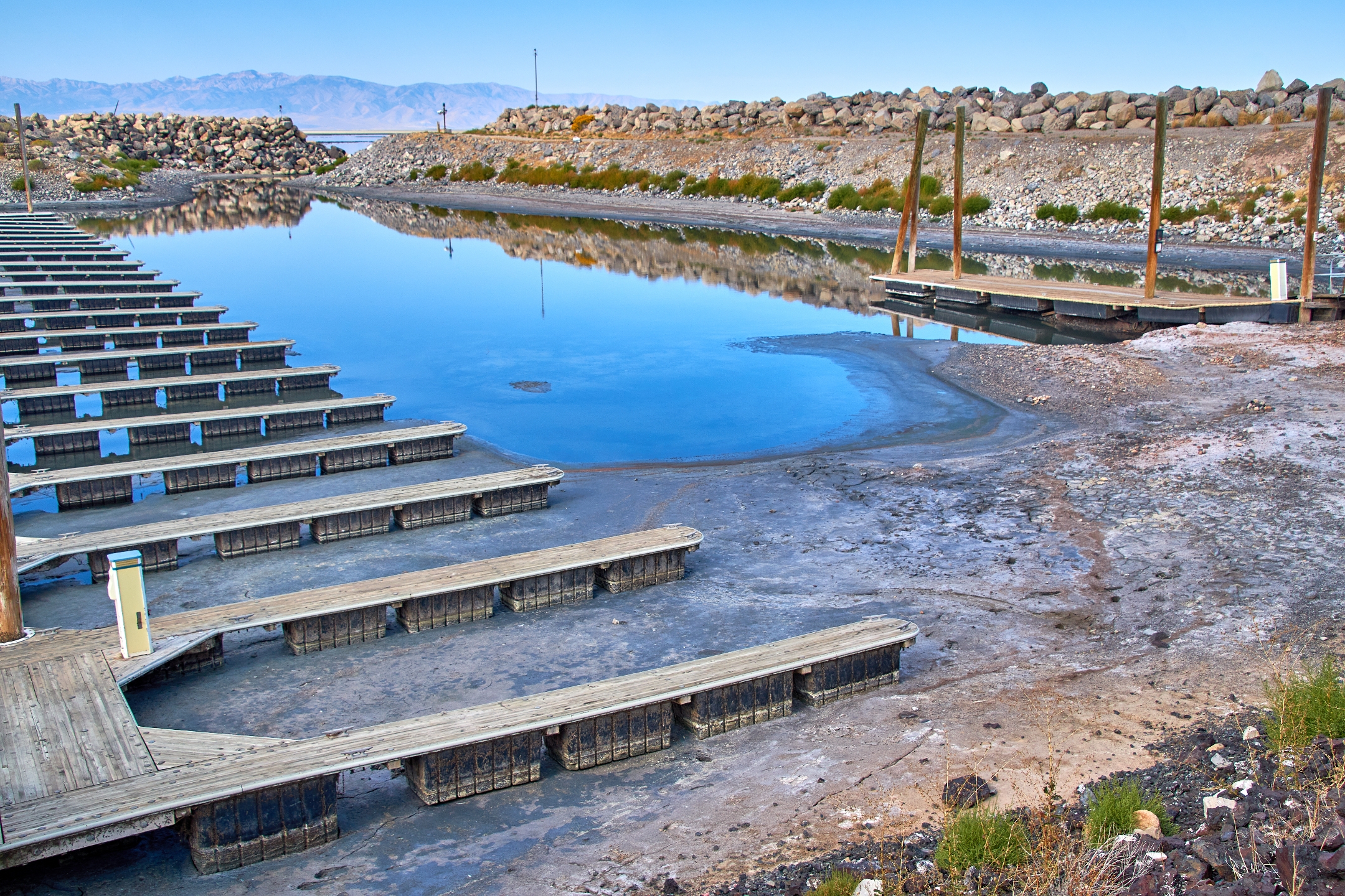 Empty wooden docks with a dry, low-water lakebed and a small body of water remaining. Rocky terrain surrounds the area