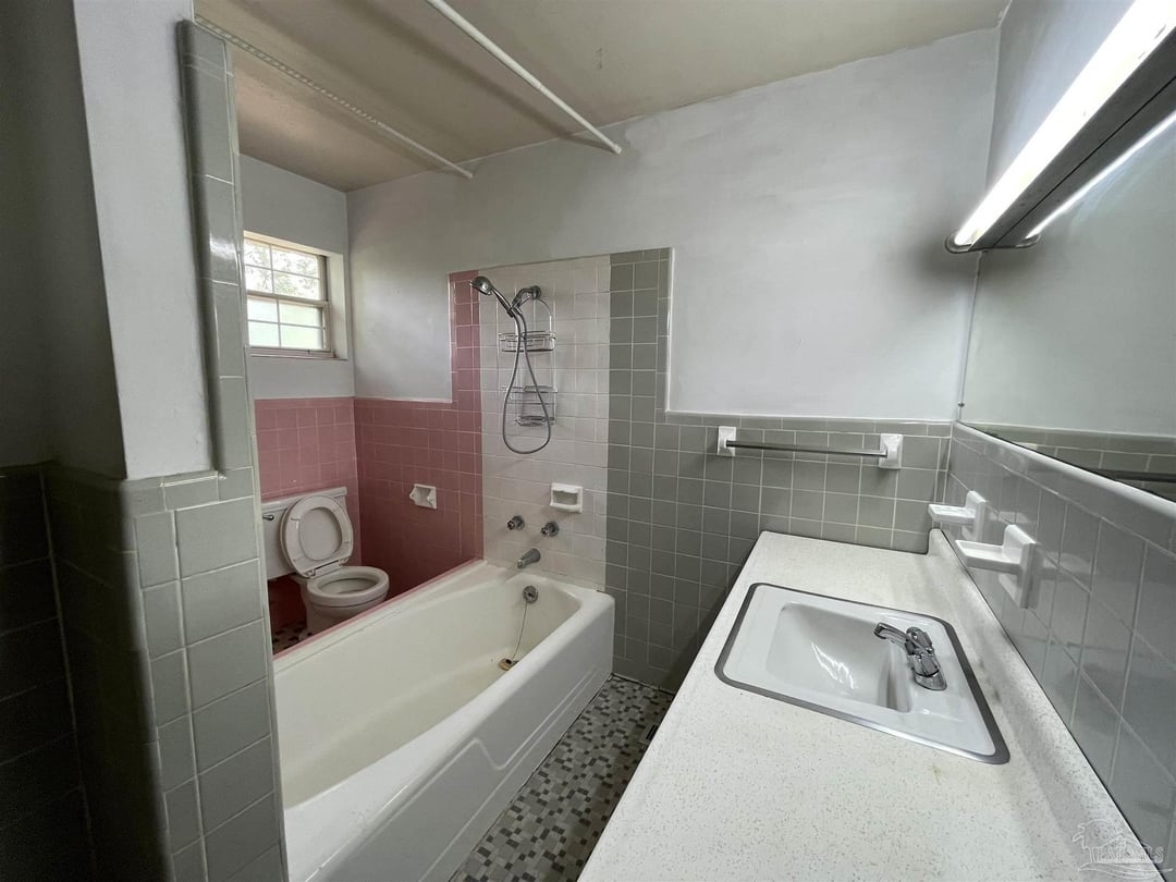 Bathroom with bathtub, shower, toilet, and sink. Walls have tile design. Natural light from a small window in the corner