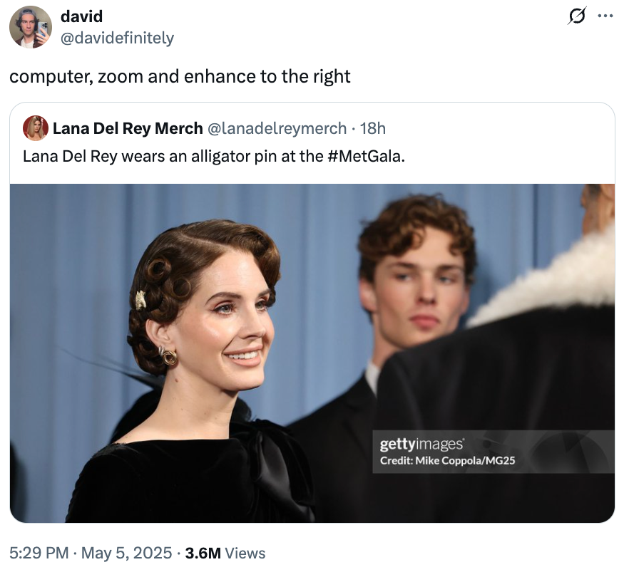 A person on a red carpet event wearing a pin in the shape of an alligator. Another individual is visible in the background