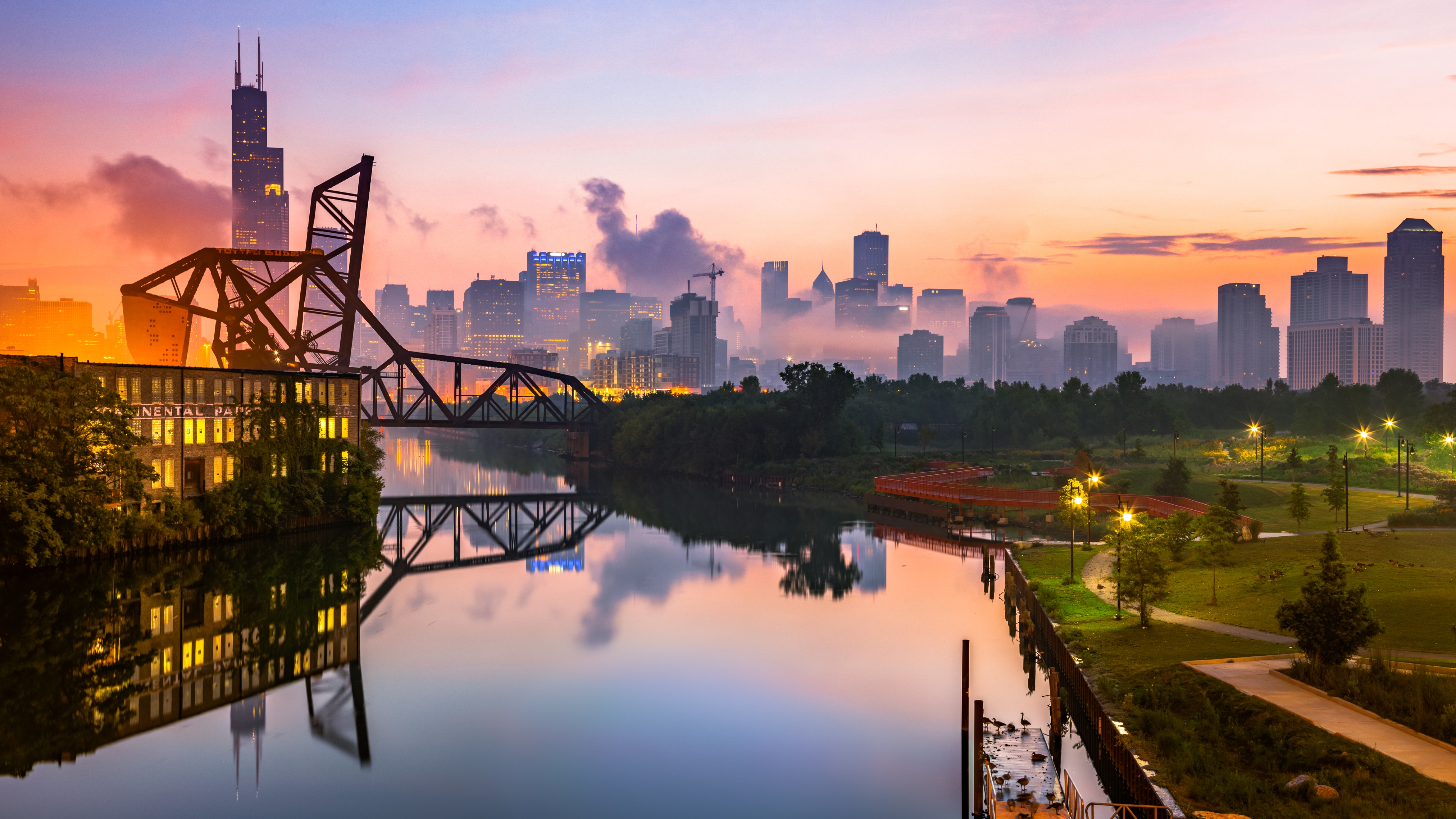 Chicago skyline at dawn with buildings reflecting in the river, bridge in foreground, and a hazy sky