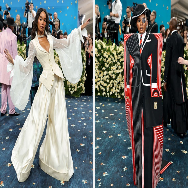 Two individuals on a red carpet: one in flowing white attire, the other in a striking abstract outfit with wide-shouldered jacket and hat