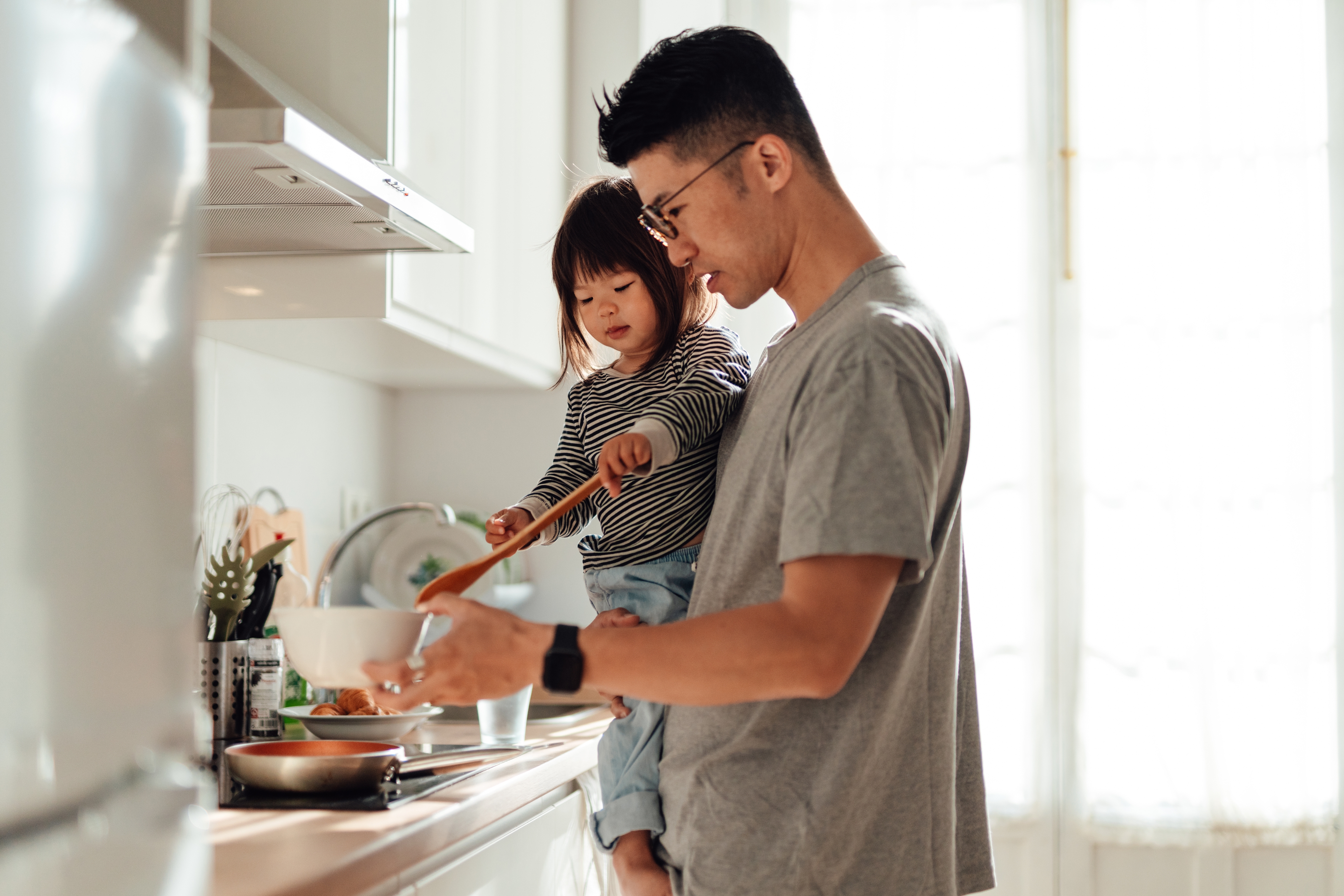 Man holding toddler helps cook in kitchen, using a wooden spoon over a bowl, while standing by a counter with food and cookware