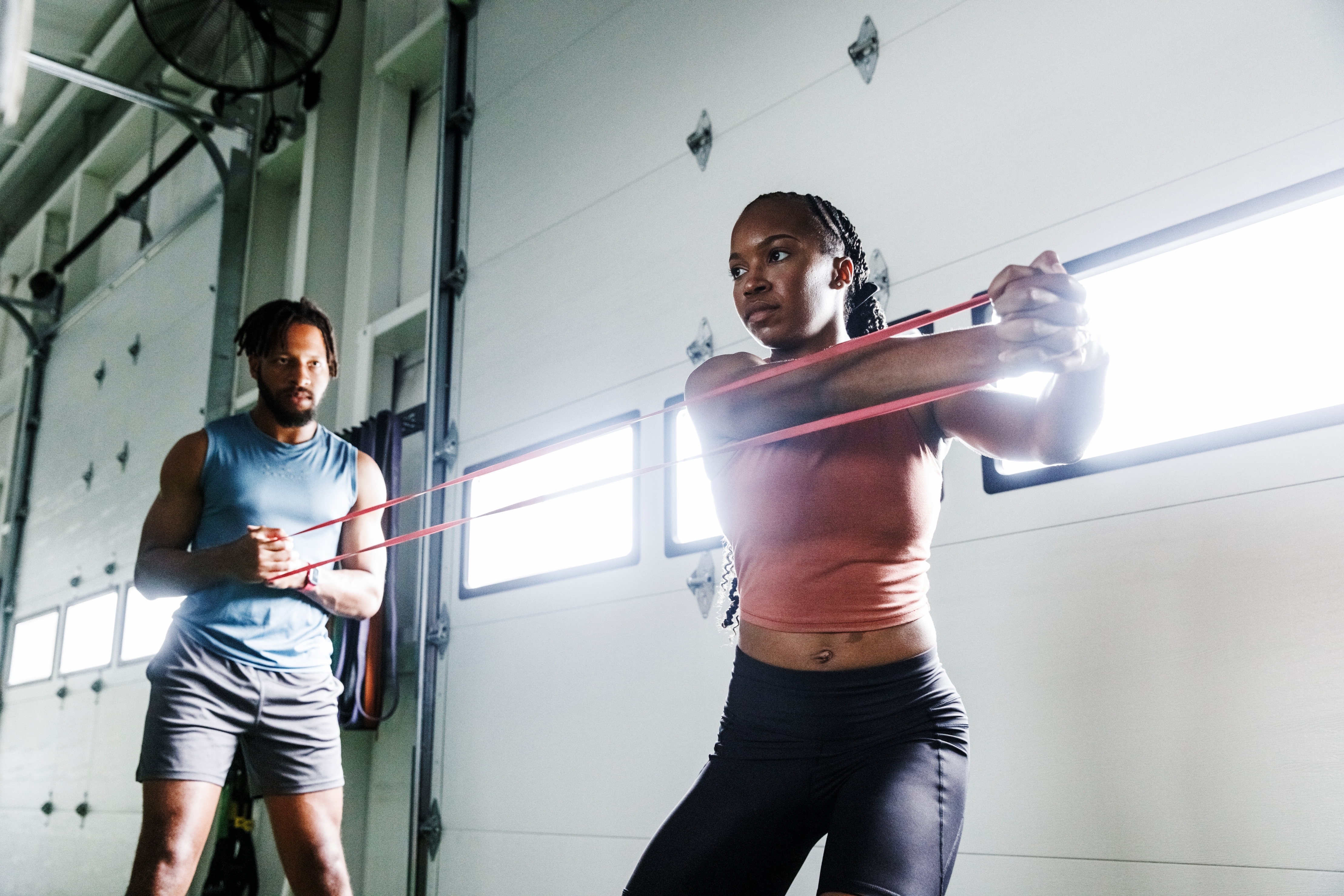 Two people in a gym, exercising with resistance bands. The woman focuses on her workout while the man observes, offering guidance
