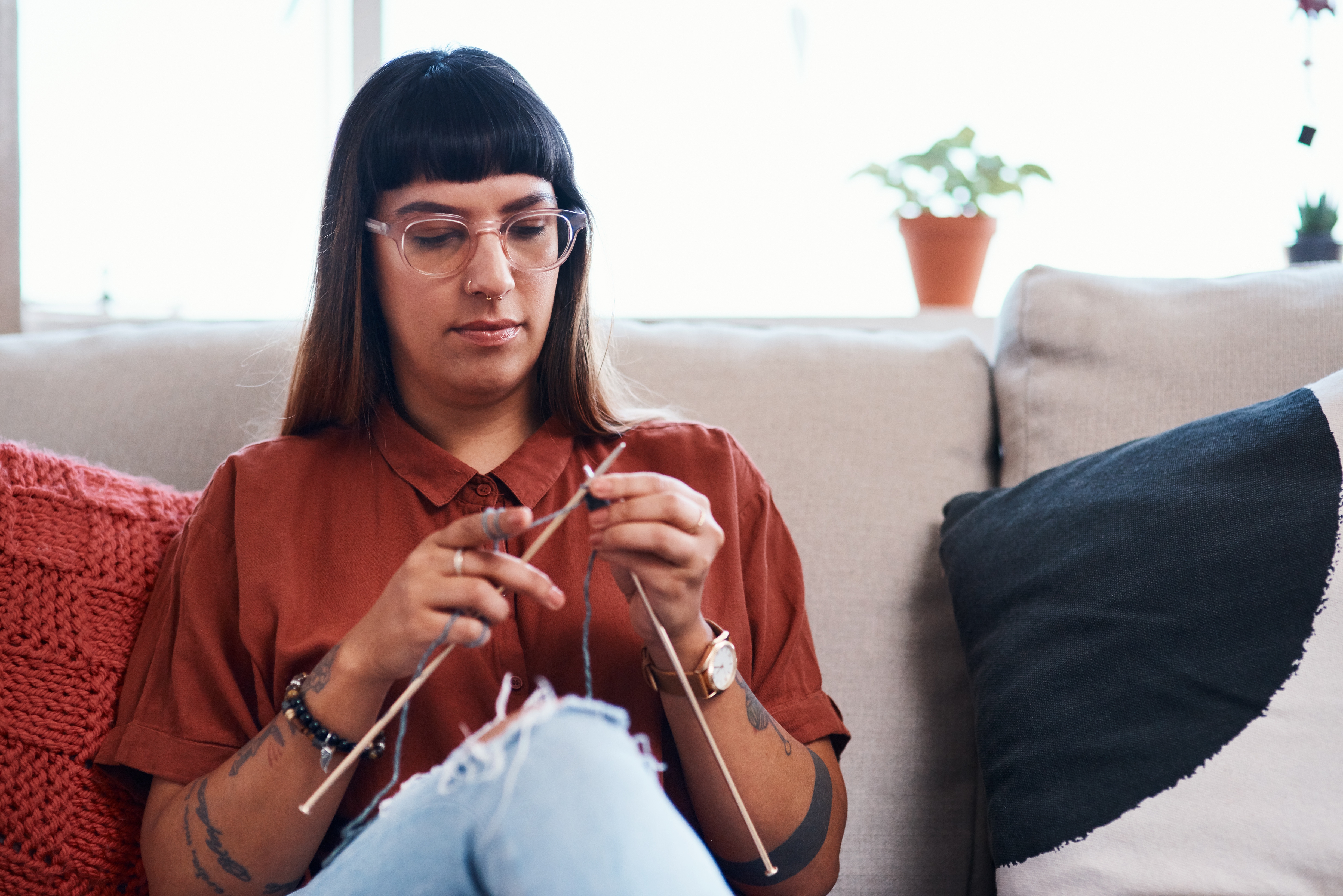 A person with glasses knits while sitting on a couch, appearing focused and relaxed