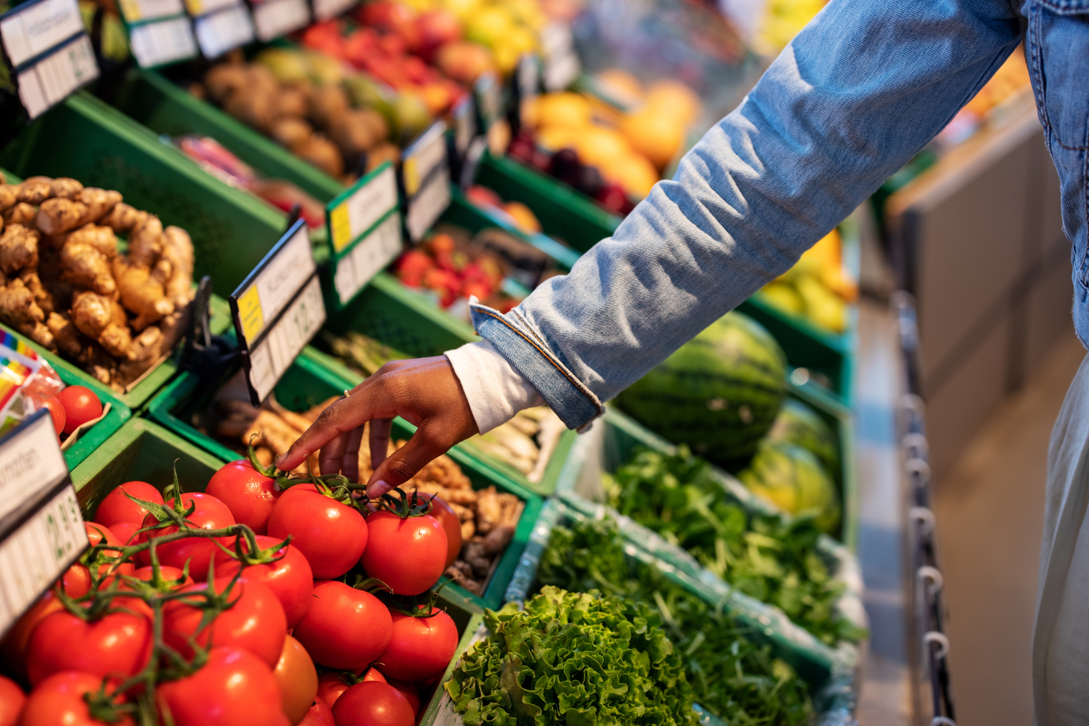 Person selecting fresh tomatoes in a grocery section with various fruits and vegetables, including greens and watermelons, visible in the background
