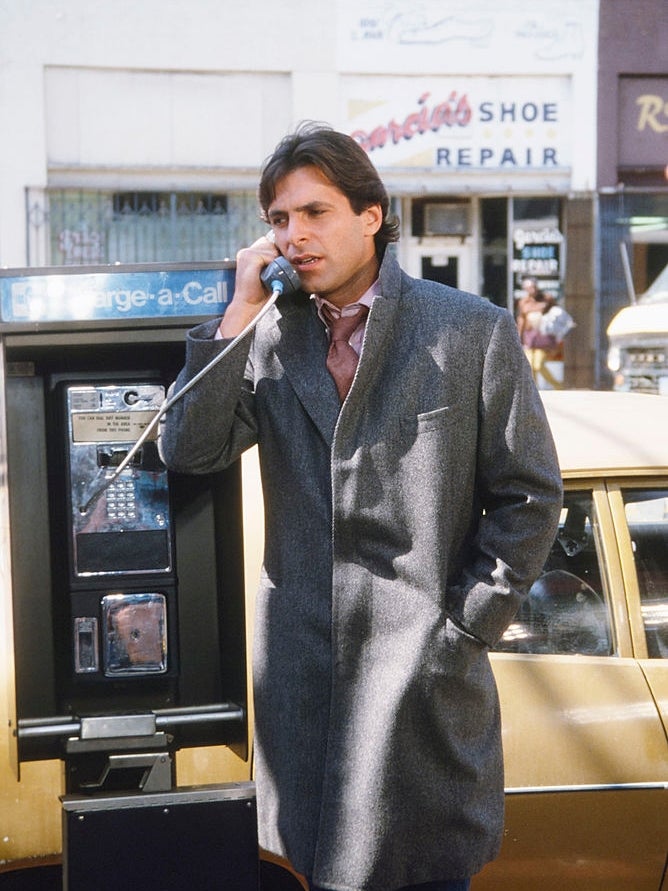 Person in a long coat uses a payphone on a city street, near a shoe repair shop and parked car