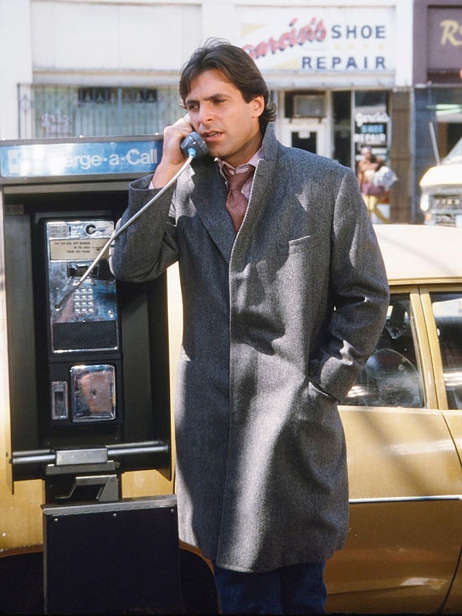 Person in a long coat uses a payphone on a city street, near a shoe repair shop and parked car