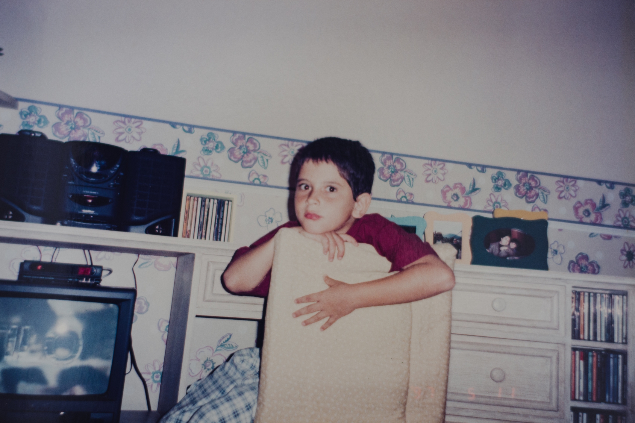 Child resting on a chair in a room with floral wallpaper, near a TV and stereo. Shelves hold CDs and framed photos