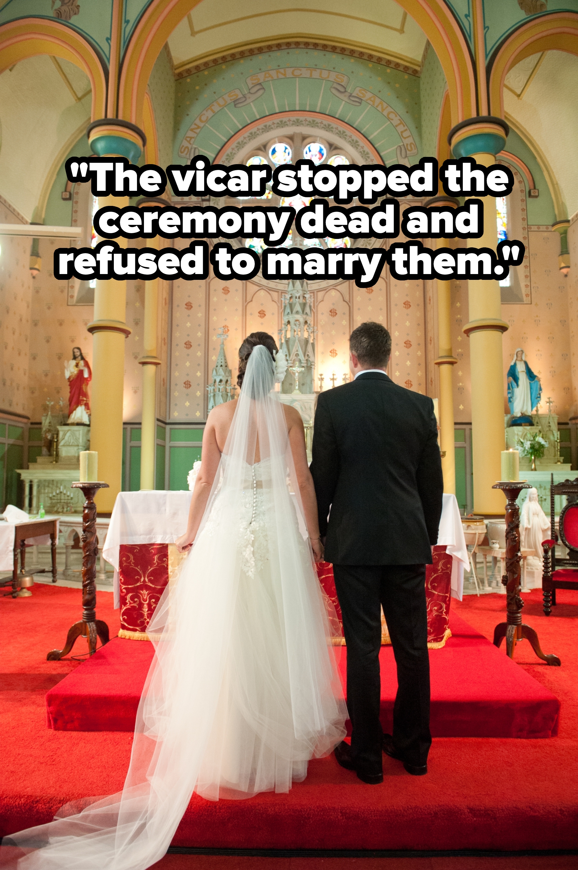 A bride in an elegant gown and veil stands beside a groom in a black suit, facing the altar in a grand church setting