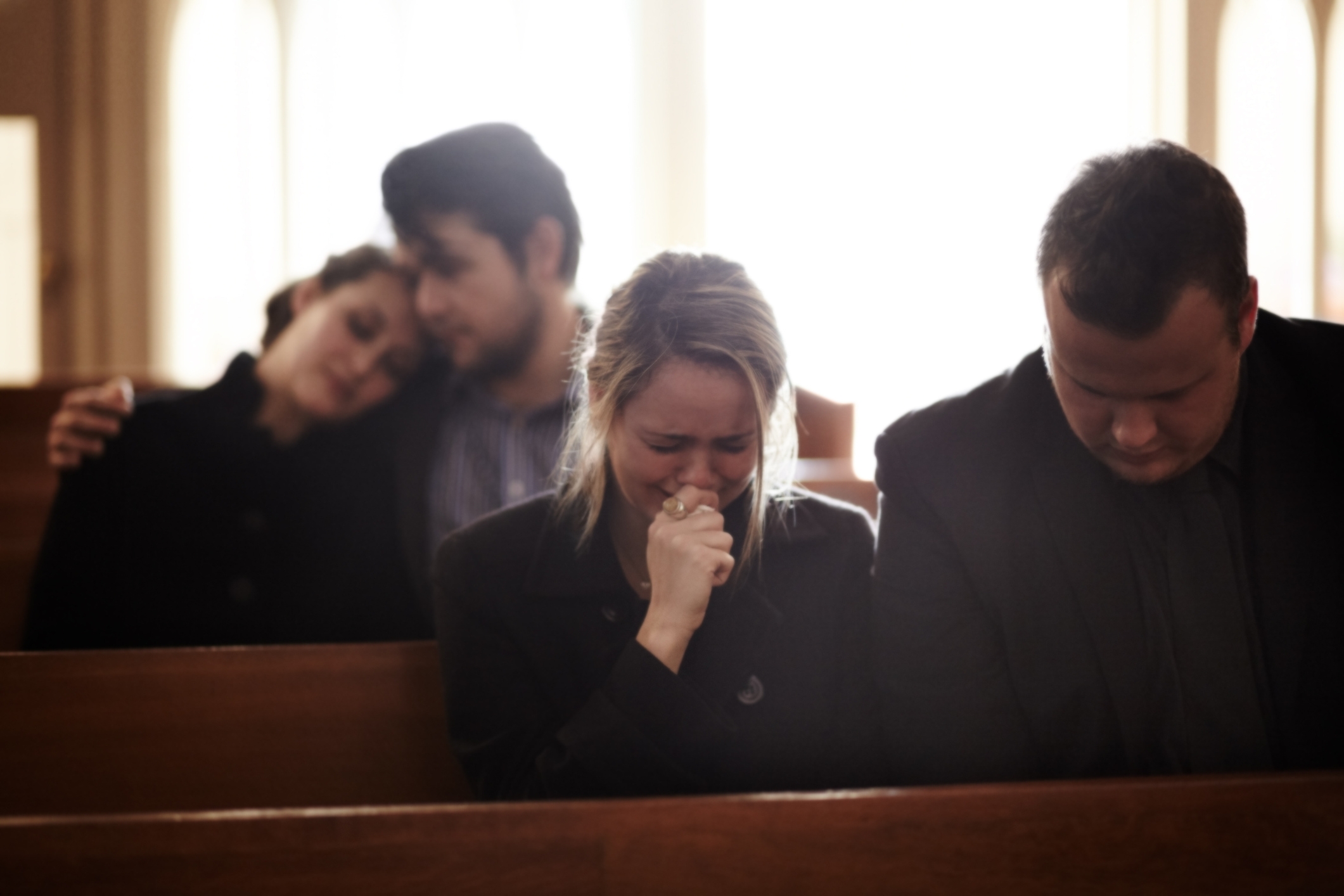 People sitting in church pews, two comforting each other, one woman visibly upset, suggesting a somber or reflective moment