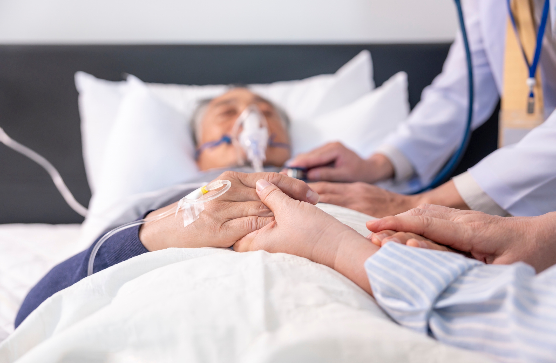 Person in hospital bed with oxygen mask, receiving care. Medical professional checks vitals while another person holds the patient’s hand for comfort