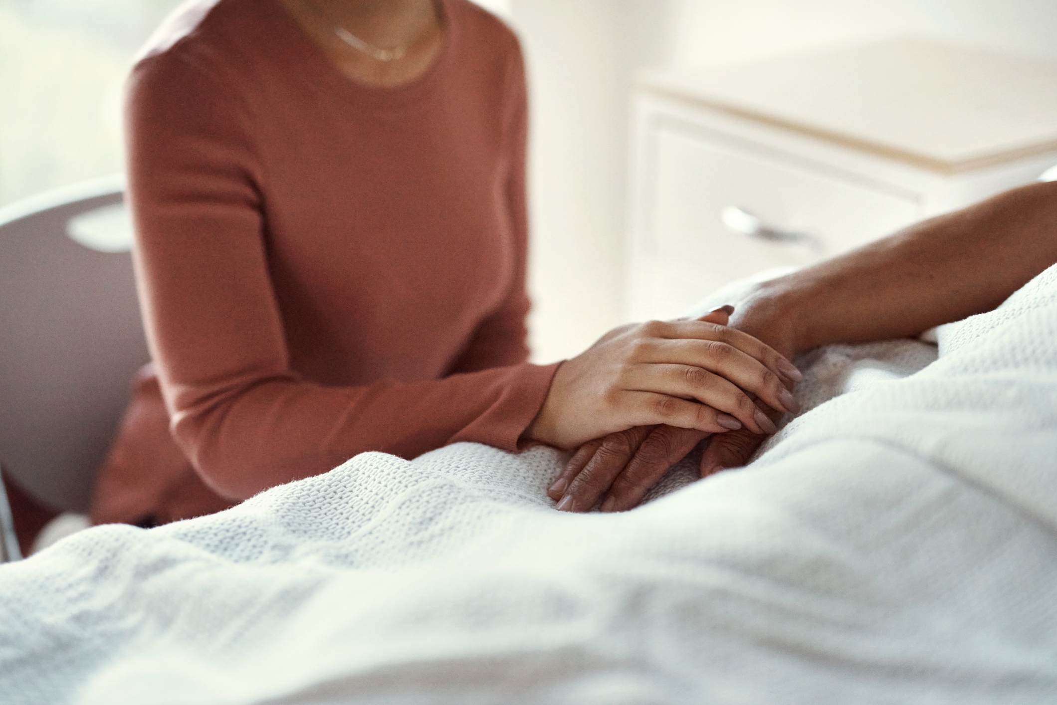 Person sitting beside a bed, gently holding the hand of someone lying under a white blanket, conveying comfort and care