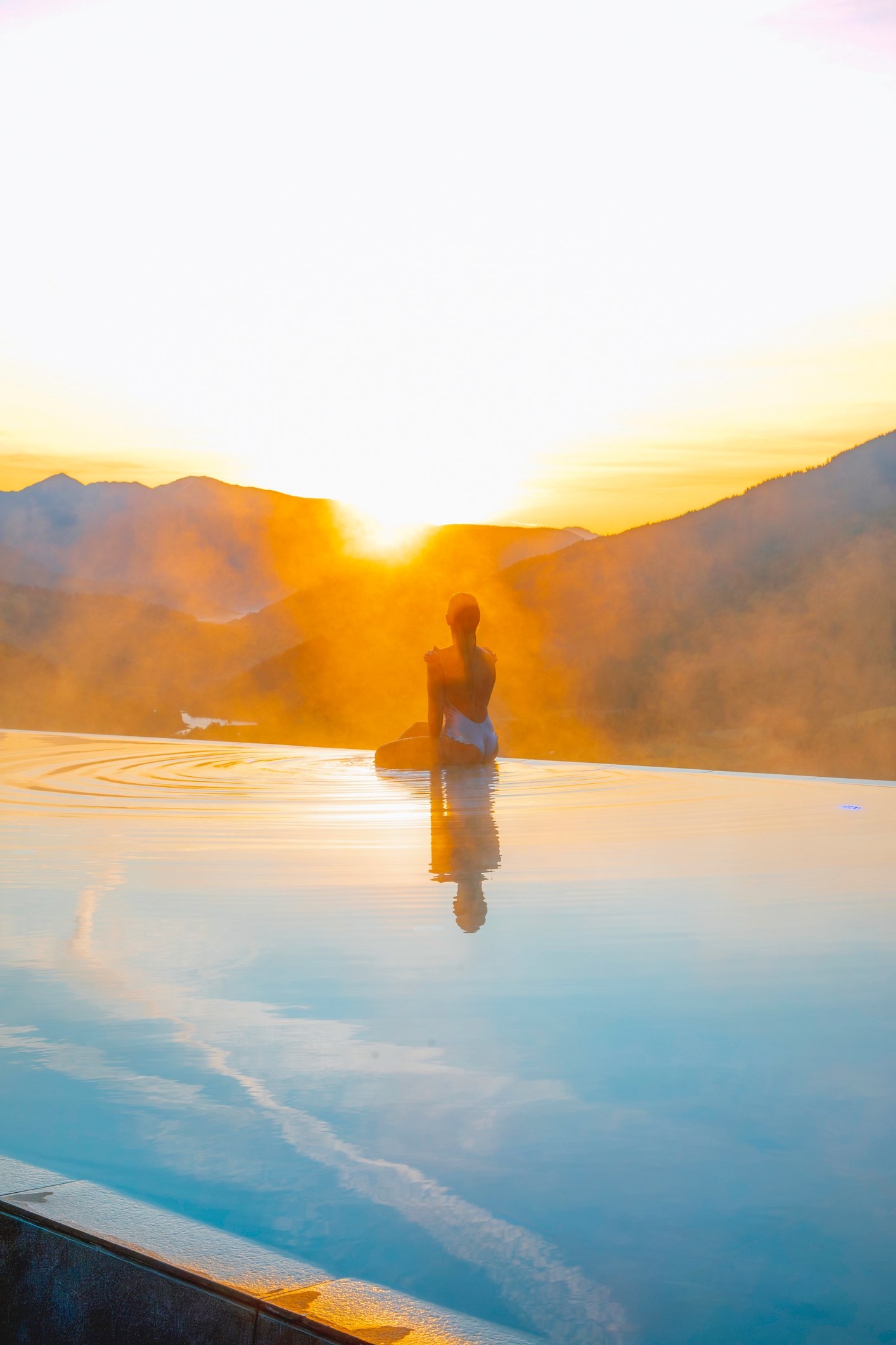 A person sits meditatively at the edge of a pool, overlooking misty mountains at sunrise